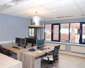 Kantoorruimte met houten bureaus, meerdere computerschermen en blauwe ramen met uitzicht op een bakstenen gebouw.