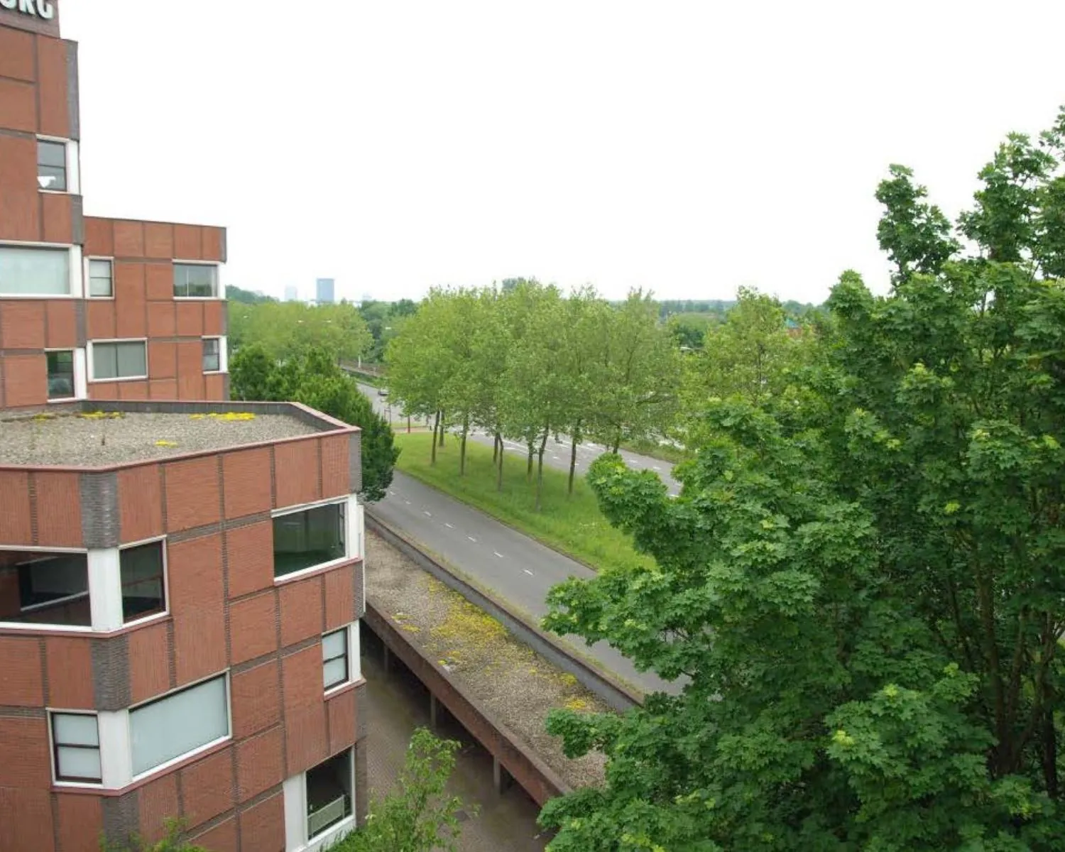 Zicht op een rood bakstenen kantoorgebouw langs een boomrijke weg met fietspad in een groene omgeving.
