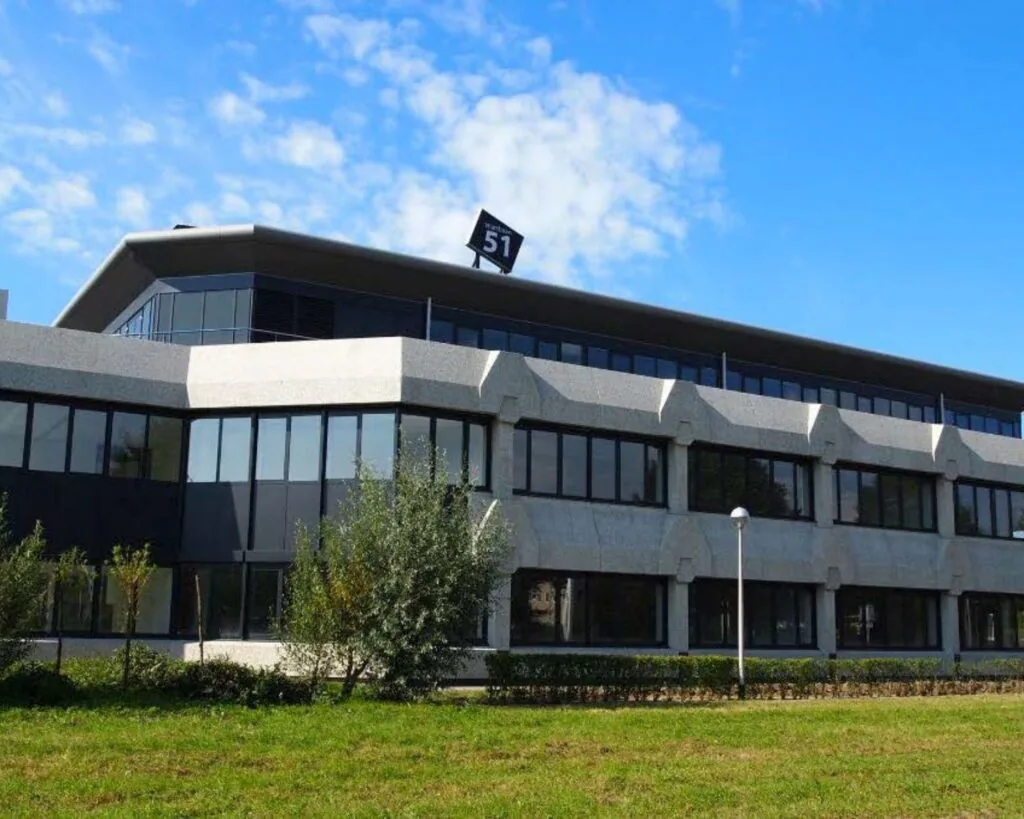 Modern kantoorgebouw met het adresnummer 51 op het dak, omgeven door groen en een blauwe lucht.