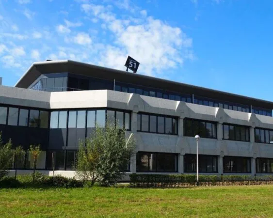 Modern kantoorgebouw met het adresnummer 51 op het dak, omgeven door groen en een blauwe lucht.