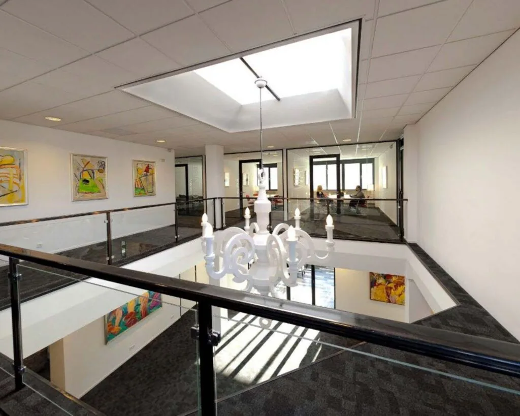 Moderne kantoorruimte met vide, glazen balustrade, kunst aan de muren en een witte kroonluchter onder een plafondraam.