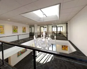 Moderne kantoorruimte met vide, glazen balustrade, kunst aan de muren en een witte kroonluchter onder een plafondraam.