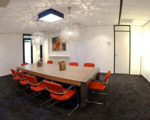 Vergaderruimte met een grote houten tafel, tien oranje stoelen en moderne hanglampen.