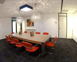 Vergaderruimte met een grote houten tafel, tien oranje stoelen en moderne hanglampen.