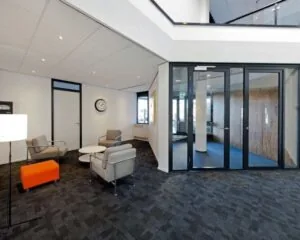 Moderne wachtruimte met grijze stoelen, een oranje poef, glazen deuren en een wandklok.