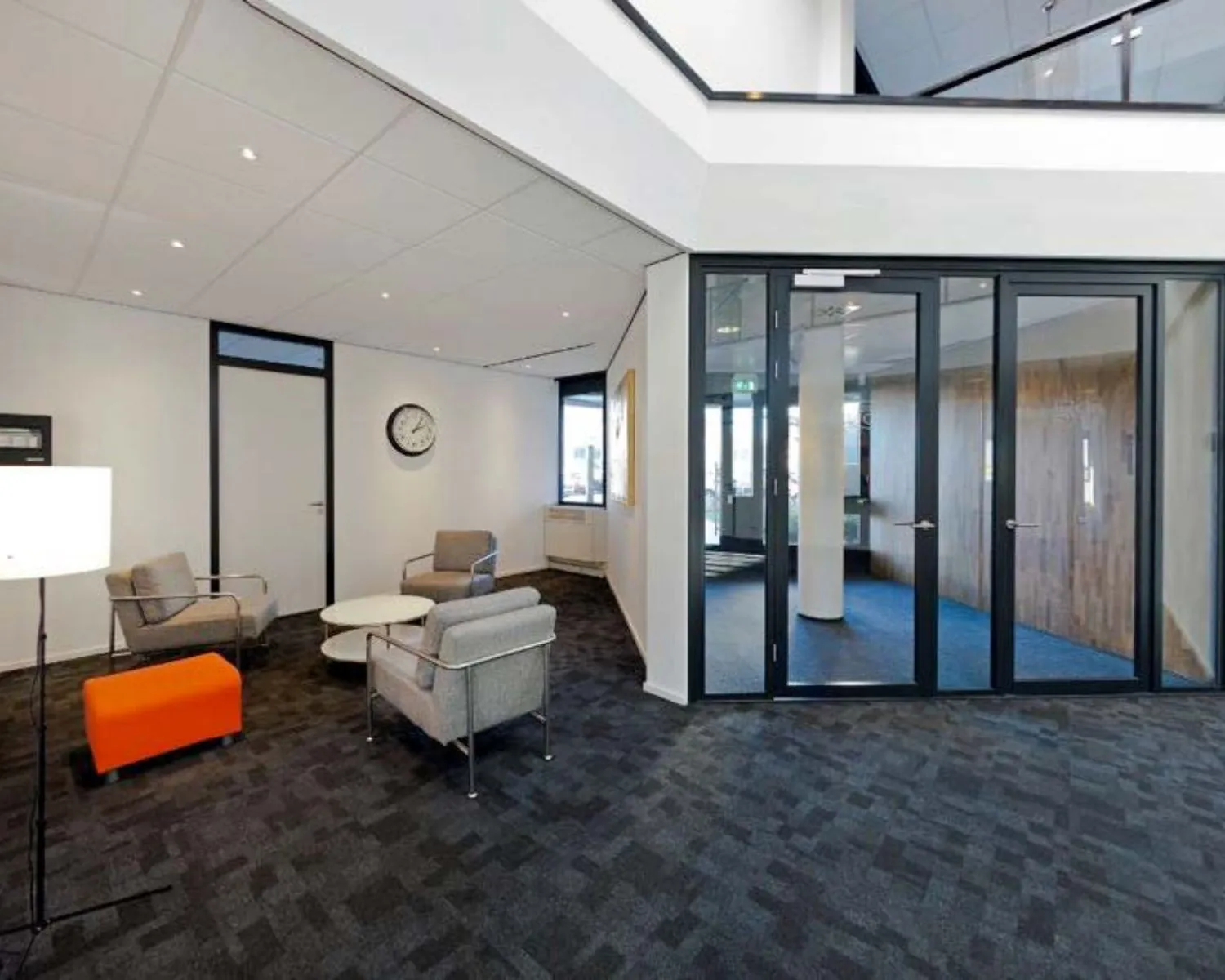 Moderne wachtruimte met grijze stoelen, een oranje poef, glazen deuren en een wandklok.
