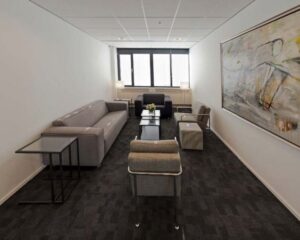 Moderne wachtruimte met grijze zitmeubelen, donker tapijt en een groot abstract schilderij aan de muur.