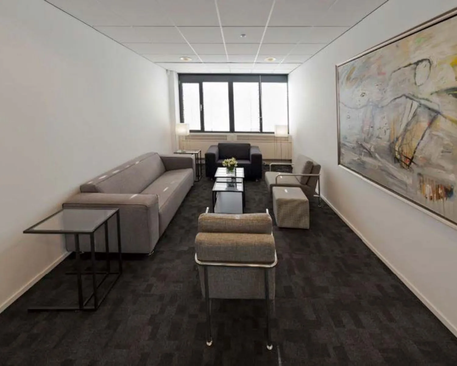 Moderne wachtruimte met grijze zitmeubelen, donker tapijt en een groot abstract schilderij aan de muur.