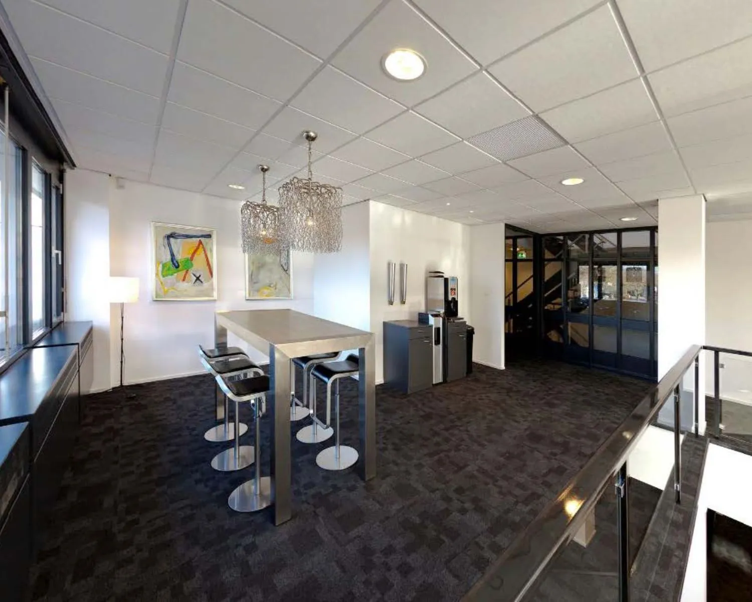 Moderne kantoorruimte met hoge tafel, barkrukken, koffiemachine en abstracte kunst aan de muur.