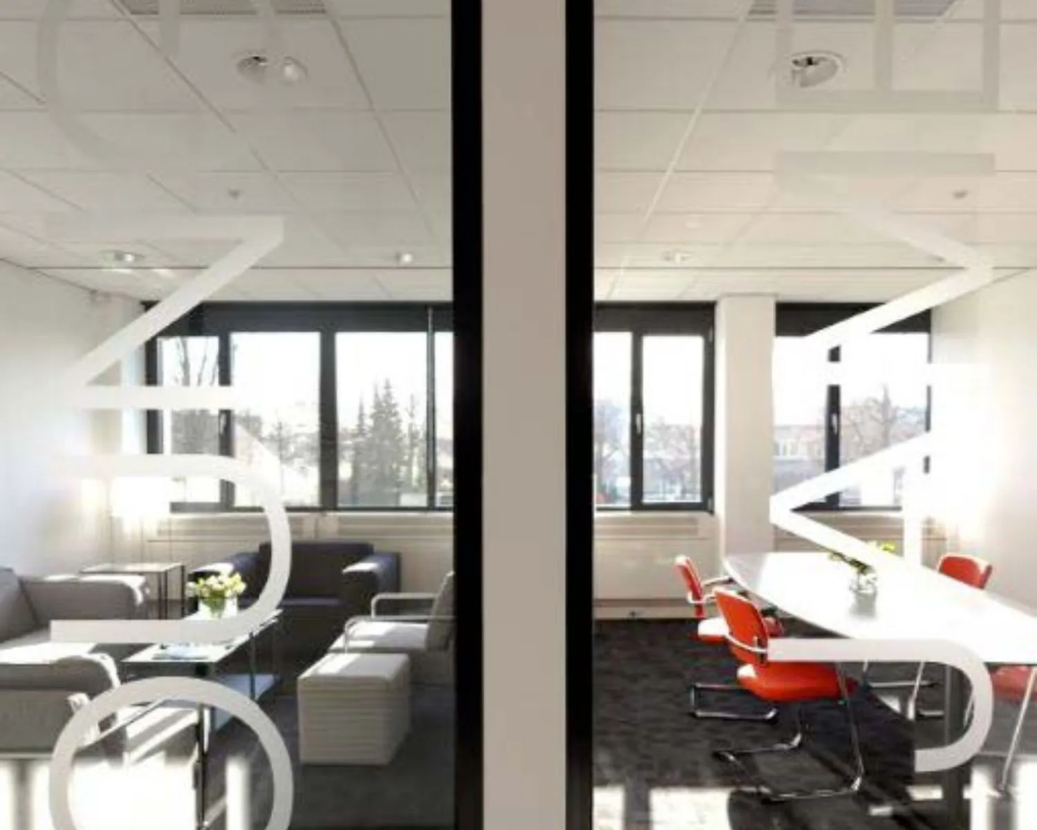 Twee moderne kantoorruimtes met glazen wand, links een zithoek en rechts een vergadertafel met rode stoelen.