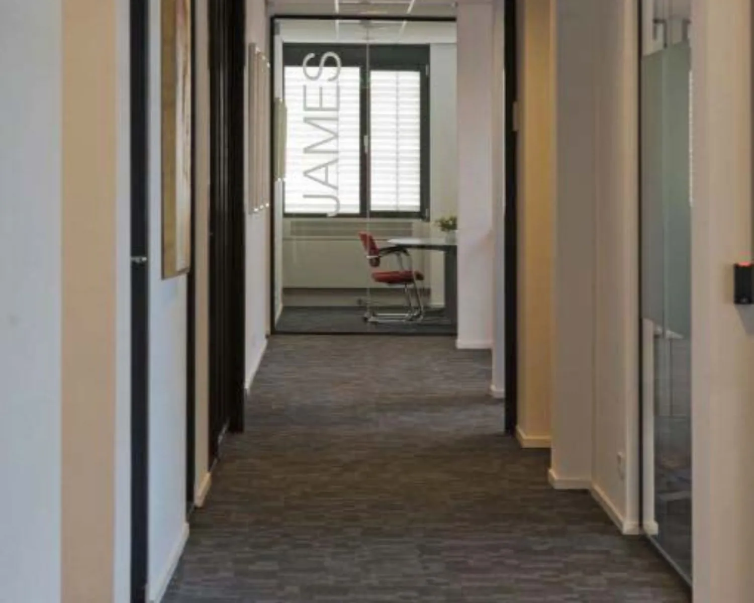 Gang in een modern kantoorpand met aan het einde een glazen vergaderruimte met de naam "JAMES" op de deur.