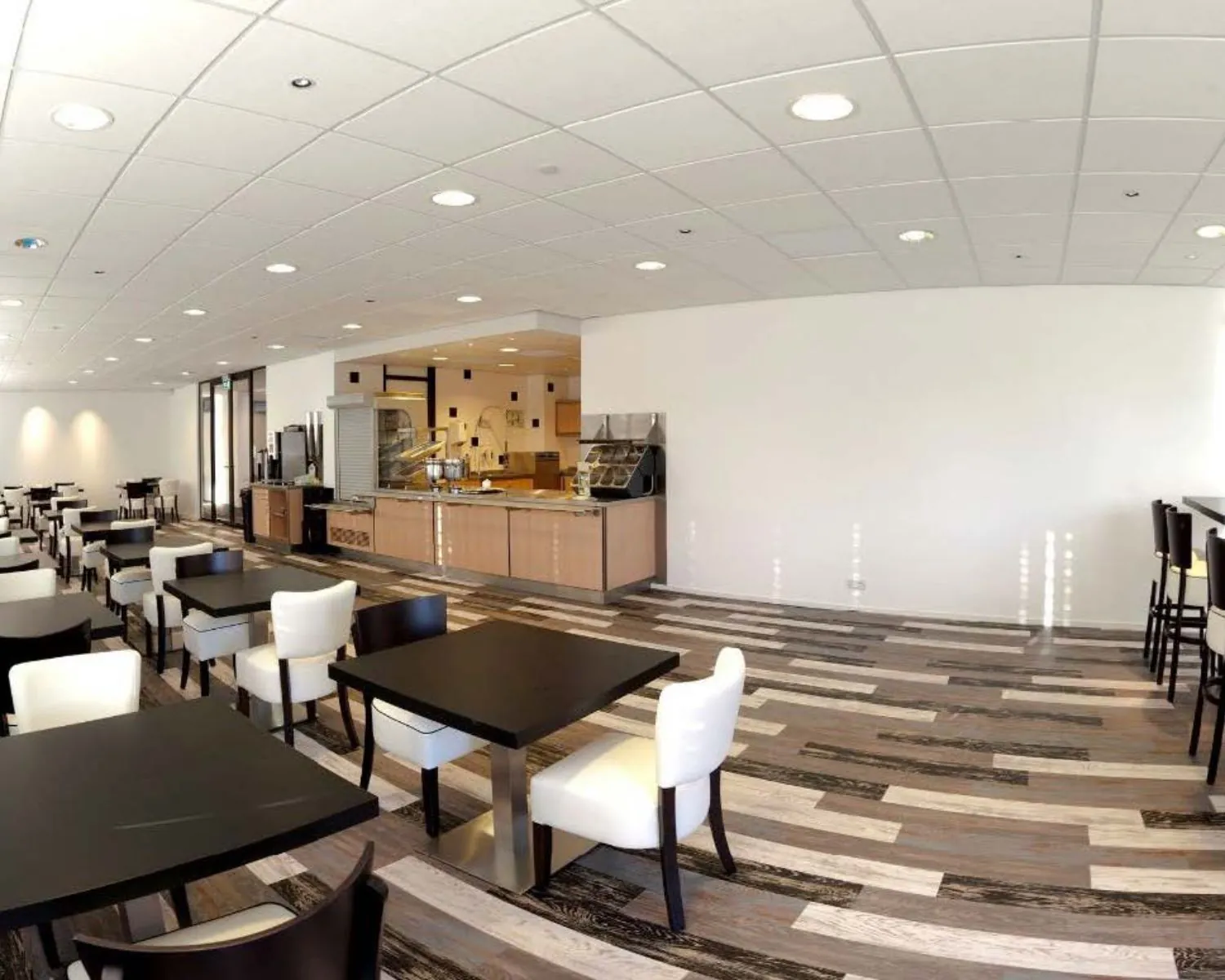 Moderne bedrijfs- of schoolkantine met zwart-witte stoelen, houten vloeren en een open buffetruimte.