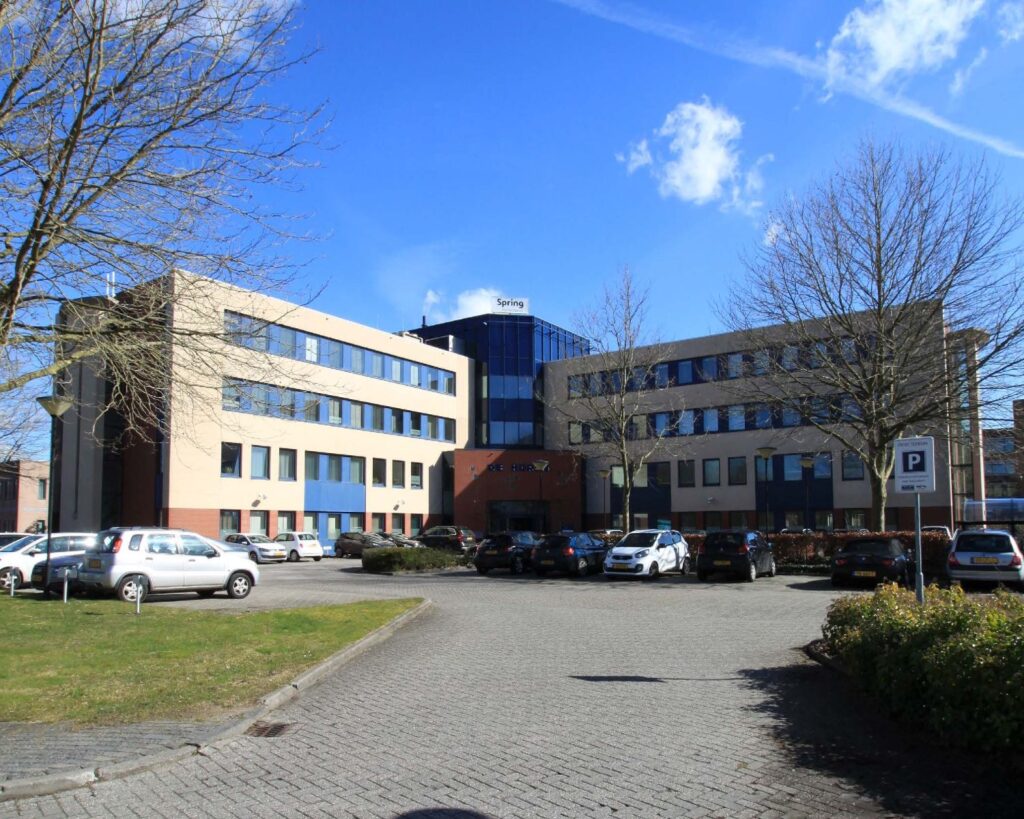 Kantoorgebouw met de naam "Spring" en een parkeerplaats op de voorgrond onder een blauwe lucht.