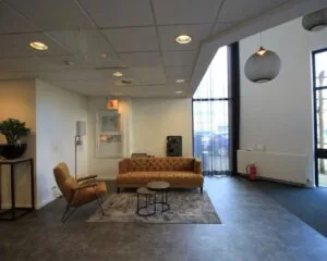 Moderne wachtruimte met een bruine bank, fauteuil, kleine salontafels en grote ramen met lichtinval.