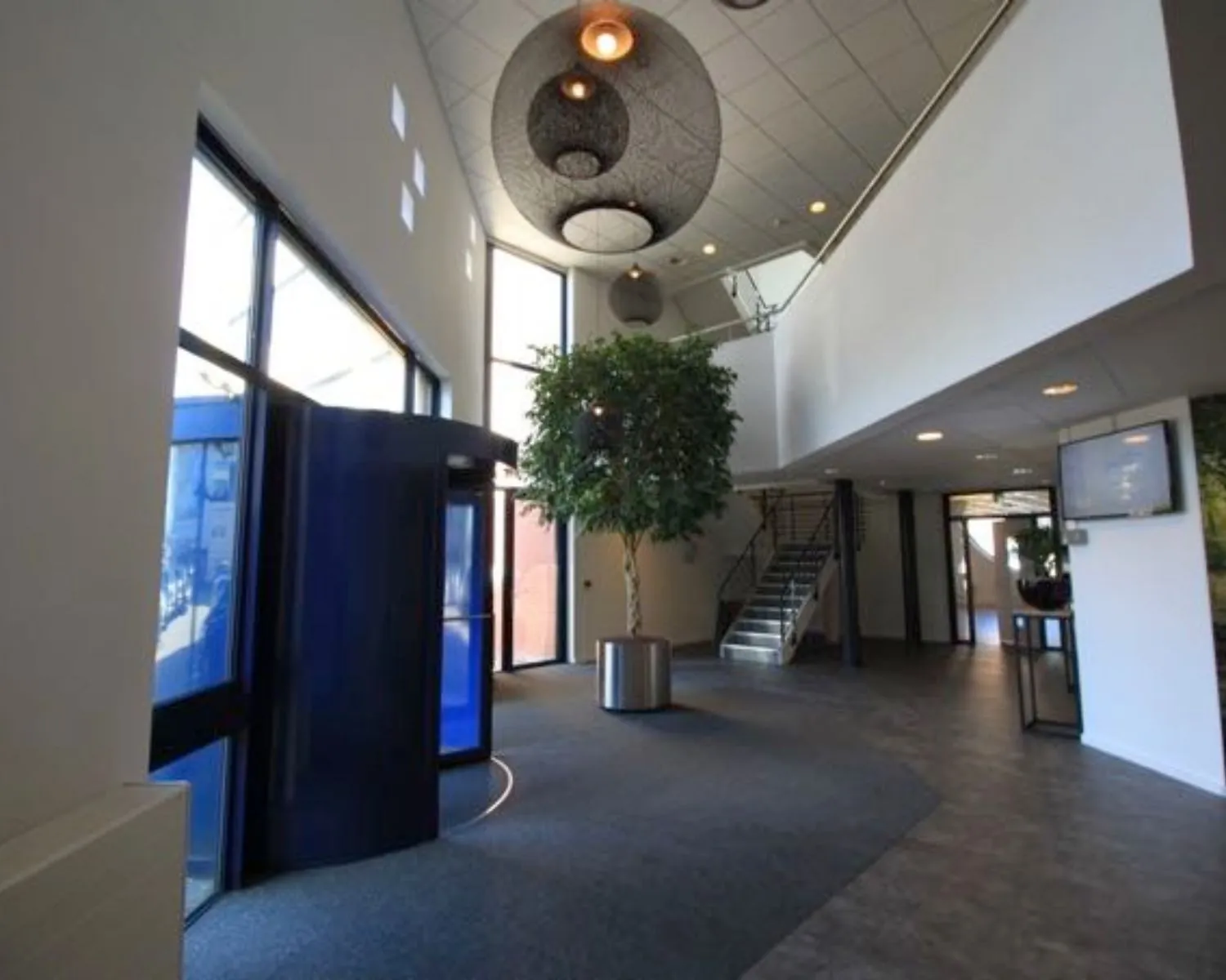 Moderne kantoorlobby met een glazen draaideur, centraal geplaatste kunstboom en een trap naar de bovenverdieping.