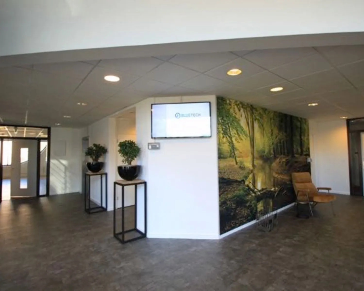 Moderne kantoorhal met houten vloer, wanddecoratie van een boslandschap, een BLUETECH-scherm en minimalistische inrichting met planten en stoelen.