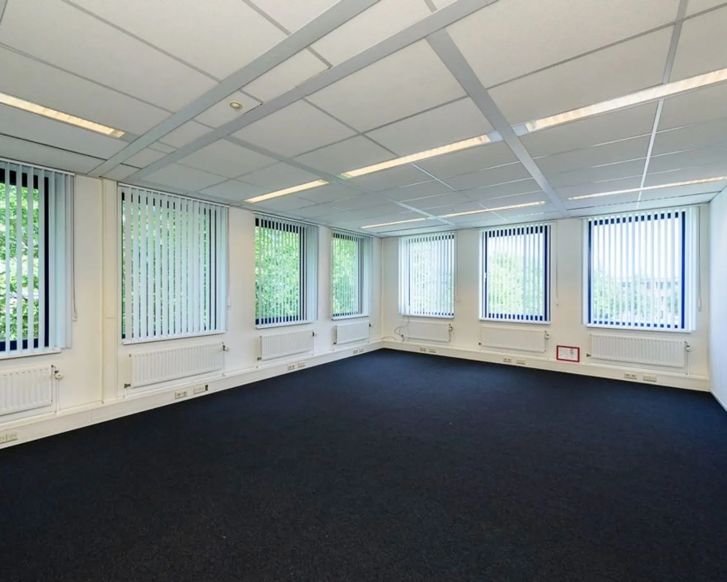 Lege kantoorruimte met donker tapijt, systeemplafond en grote ramen met verticale jaloezieën.
