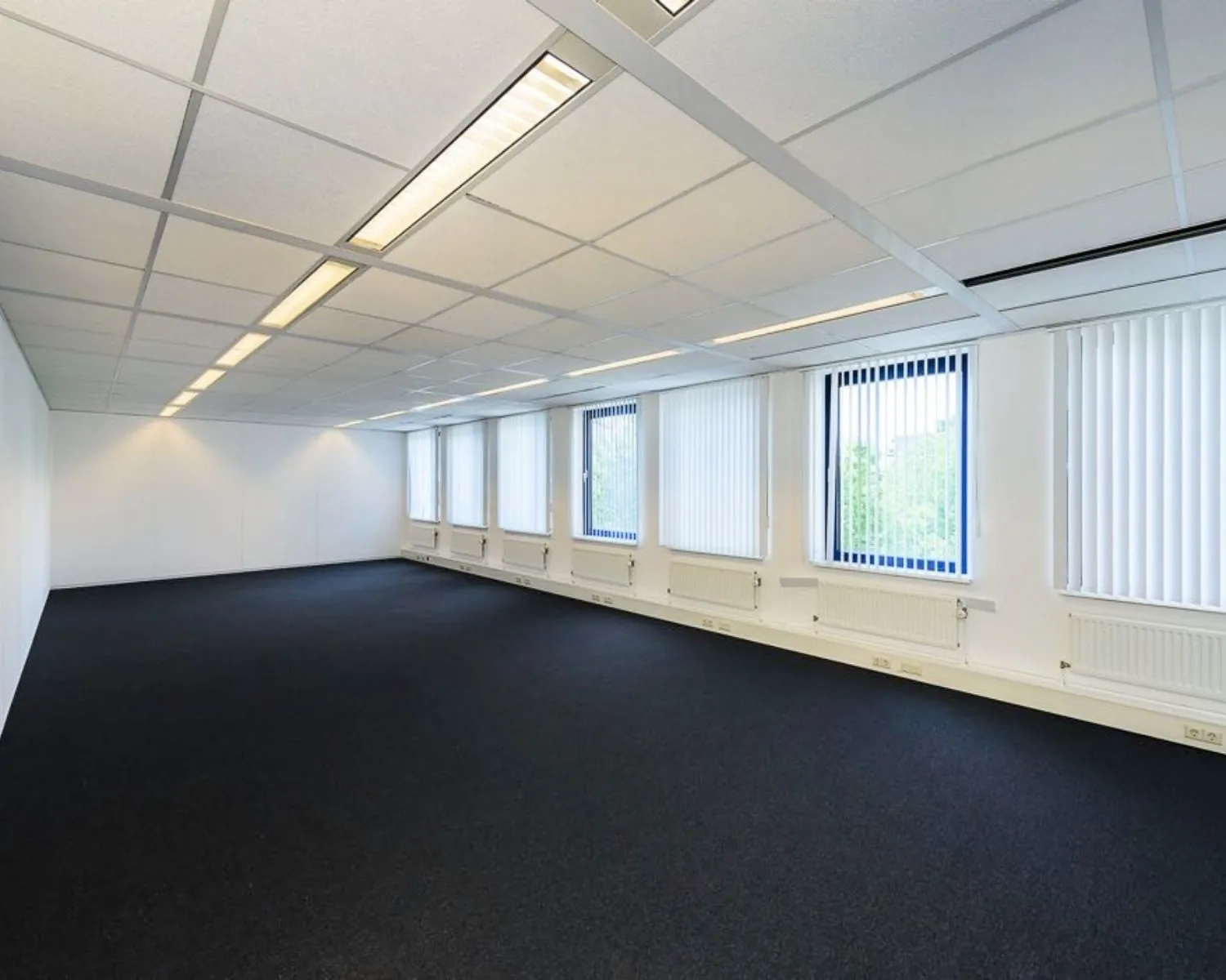 Lege, ruime kantoorruimte met donker tapijt, systeemplafond en ramen met verticale jaloezieën.