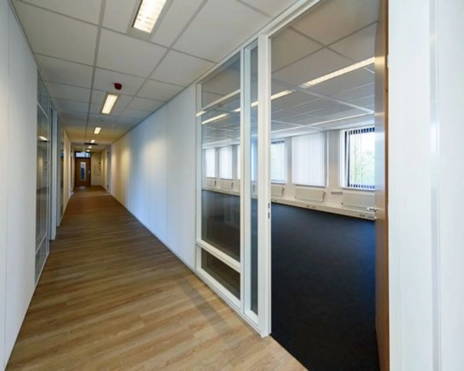 Lichte kantoorruimte met groot raam en glazen schuifdeur naast een lange gang met houten vloer.