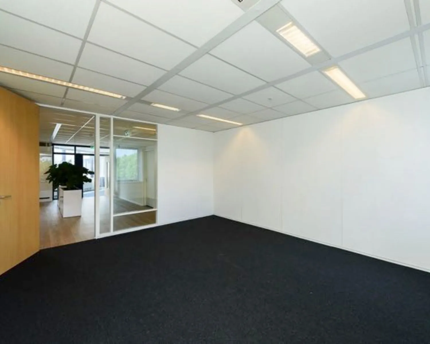 Lege kantoorruimte met witte wanden, systeemplafond en donker tapijt, grenzend aan een gang met glazen wand en houten vloer.