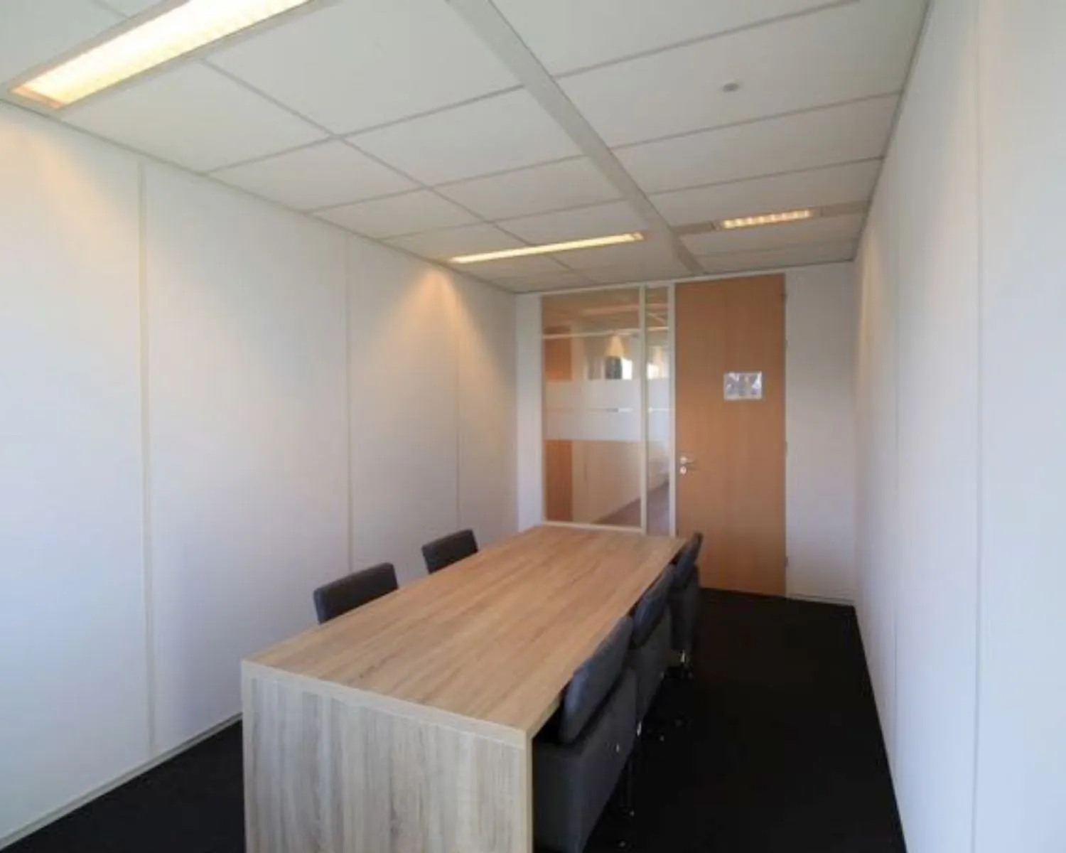 Kleine vergaderruimte met een houten tafel, zes stoelen en witte wanden.