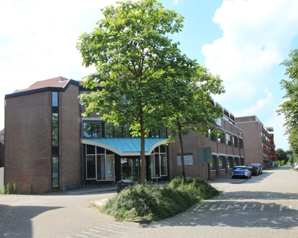 Een modern bakstenen kantoorgebouw met blauwe luifel en omliggende parkeerplaatsen op een zonnige dag.