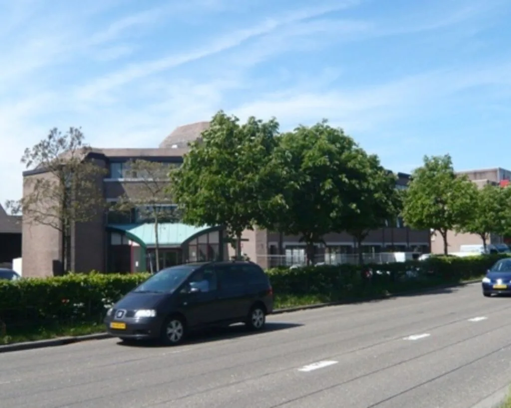 Bakstenen kantoorgebouw met grote ramen en een groene entree, gelegen aan een straat met geparkeerde auto’s en bomen.