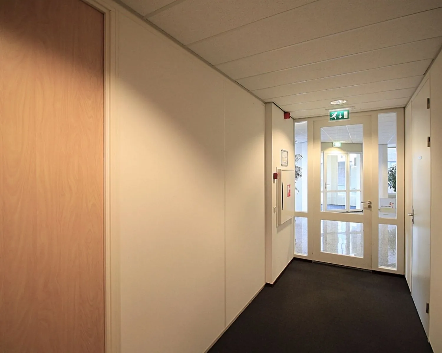 Lichte gang in een kantoorgebouw met witte muren, een zwart vloerbedekking en een glazen deur met nooduitgangbord.