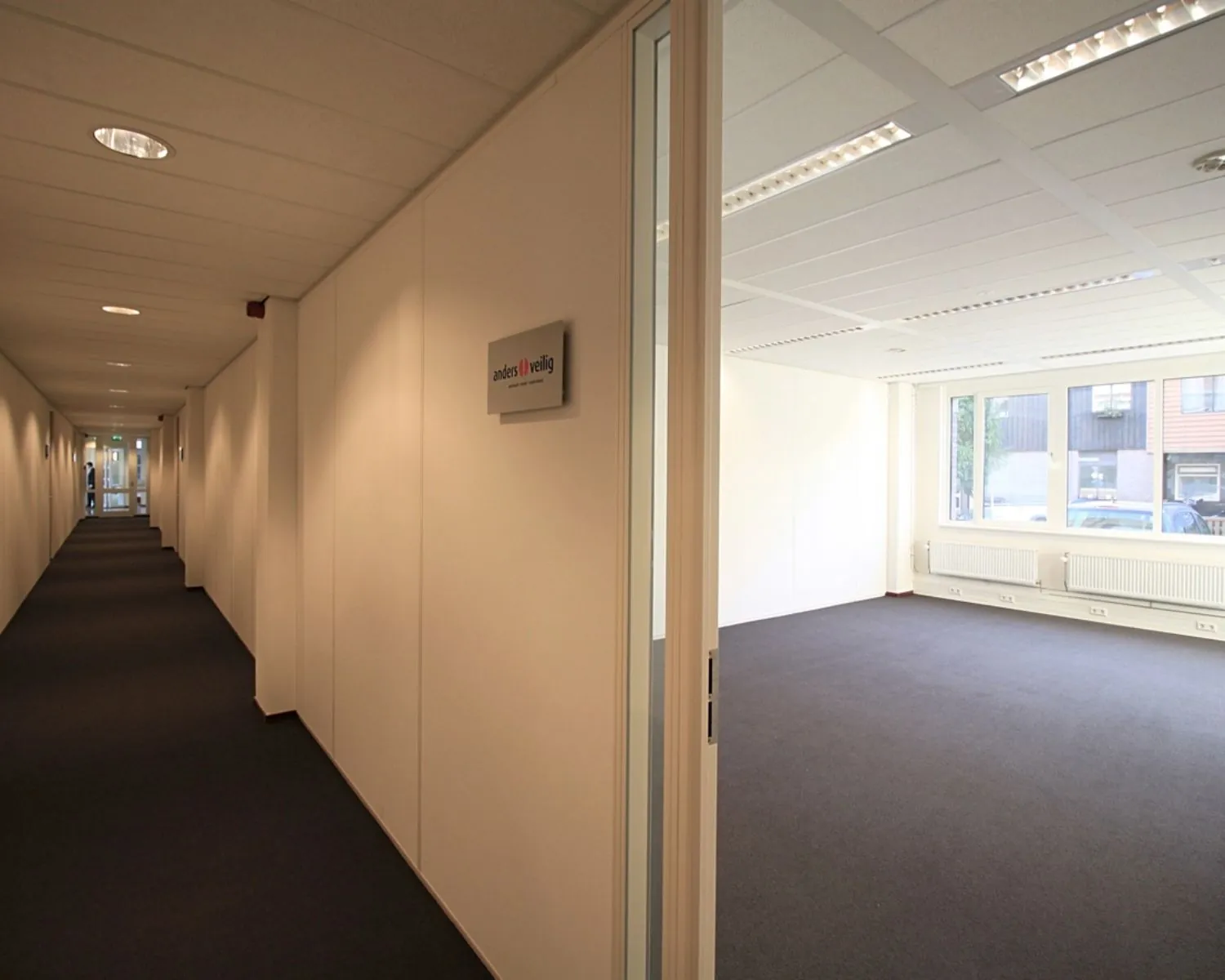 Verlichte kantoorruimte met open deur aan een lange gang met het bordje "anders // veilig" aan de muur.