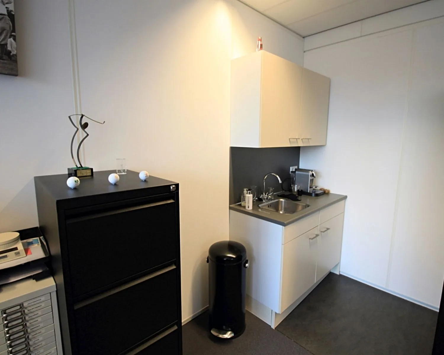 Kleine kantoorpantry met een aanrecht, koffiezetapparaat, zwarte prullenbak en ladekast met decoratie.