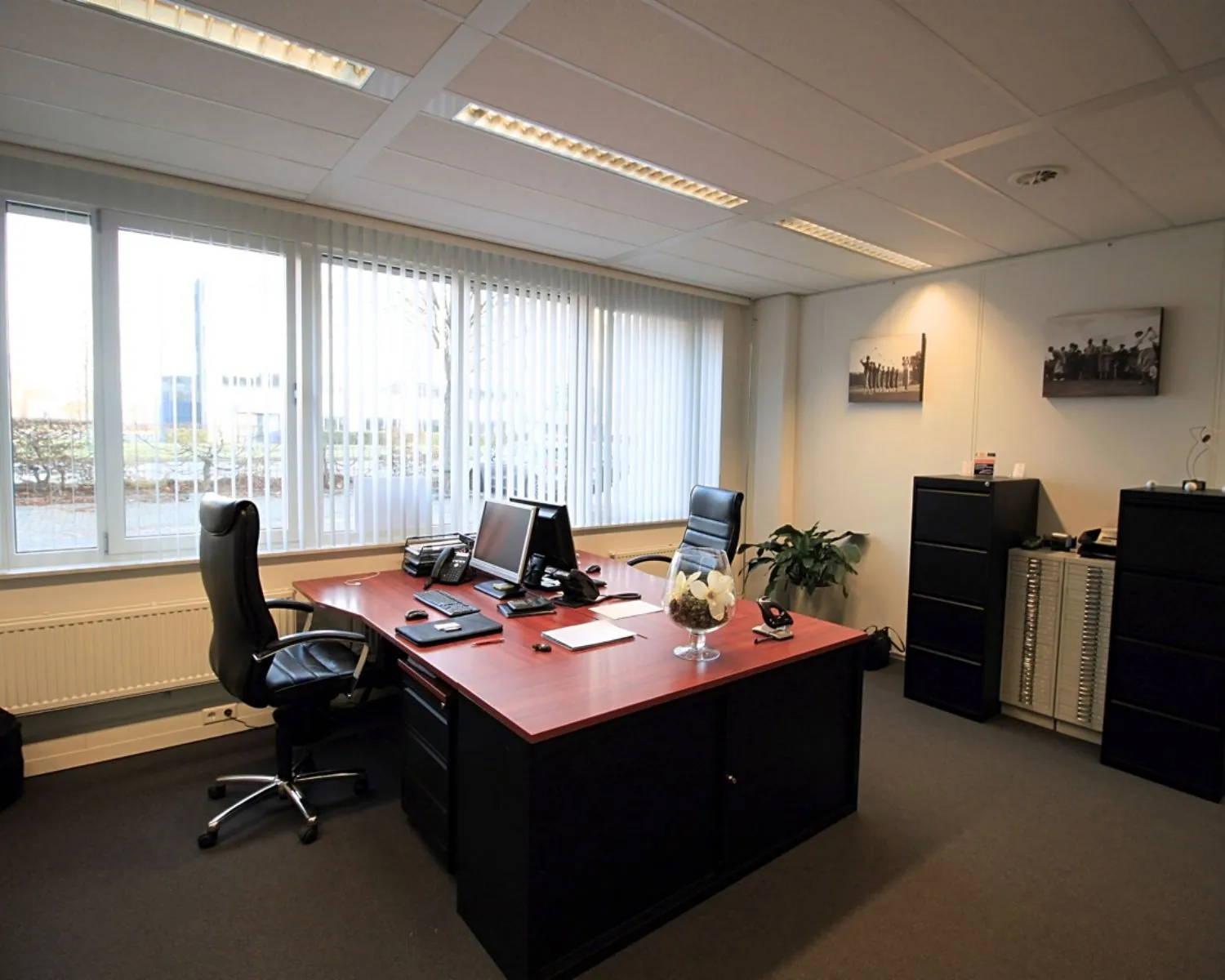 Modern ingericht kantoor met een groot bureau, twee bureaustoelen, computers en kasten, gelegen aan een raam met verticale jaloezieën.