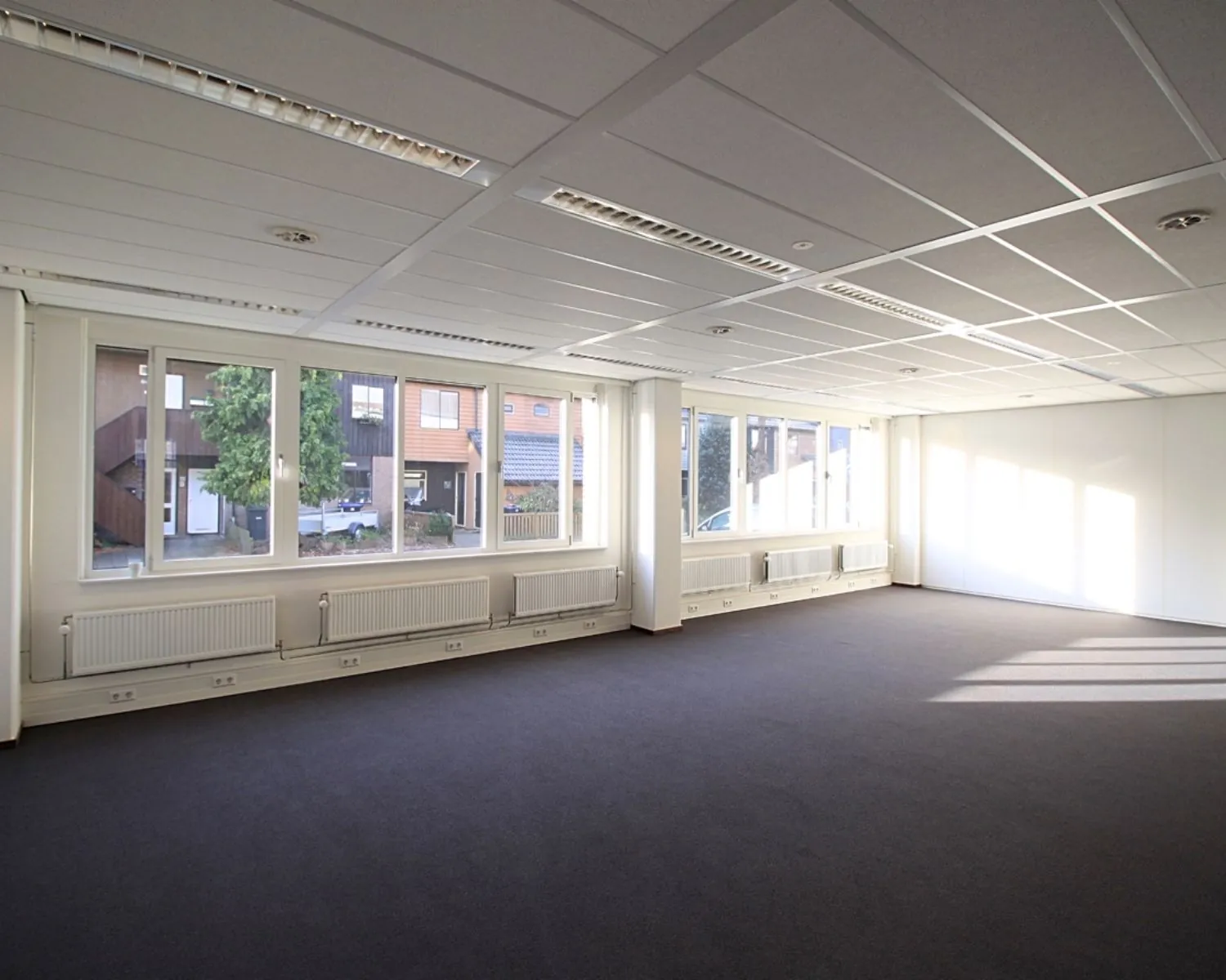 Lichte, lege kantoorruimte met grote ramen, systeemplafond en donkertapijt.