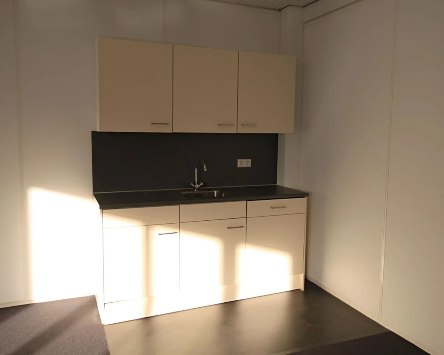 Kleine kitchenette met beige kastjes, zwart aanrechtblad en gootsteen in een minimalistisch interieur.