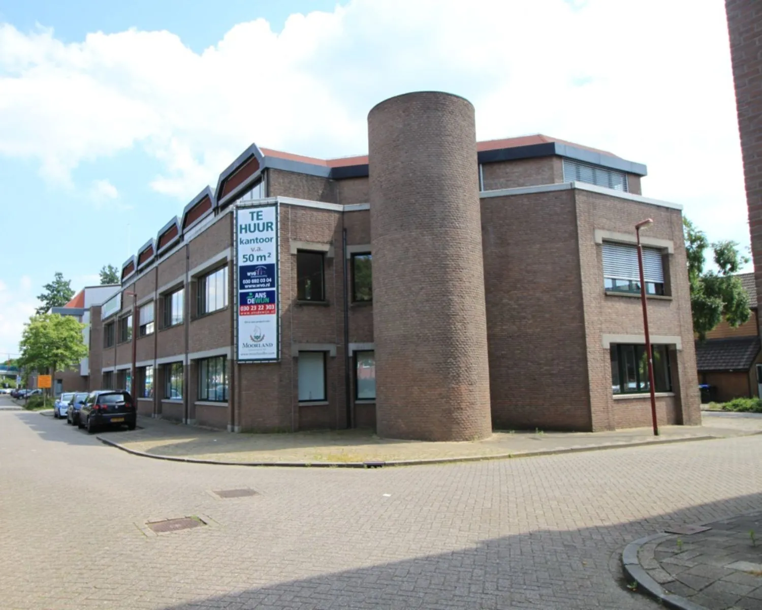 Bakstenen kantoorpand op een hoek met een bord “Te Huur” en ronde torenstructuur aan de voorzijde.
