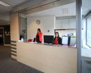 Twee receptionistes werken achter een moderne balie in een kantoorruimte met neutrale kleuren.