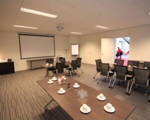 Vergaderruimte met opgestelde stoelen, tafels met koffiekopjes, projector en flip-over bij een open deur.