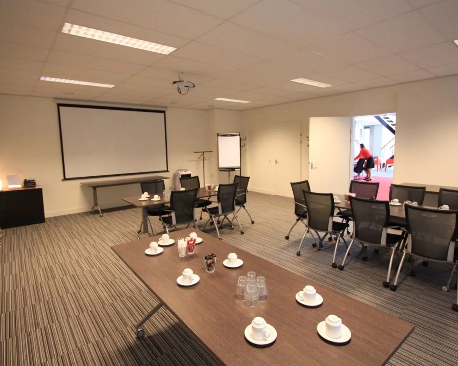 Vergaderruimte met opgestelde stoelen, tafels met koffiekopjes, projector en flip-over bij een open deur.