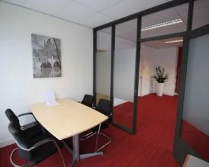 Kleine vergaderruimte met vier stoelen, een houten tafel en een zwart-wit foto aan de muur op roodgestreept tapijt.