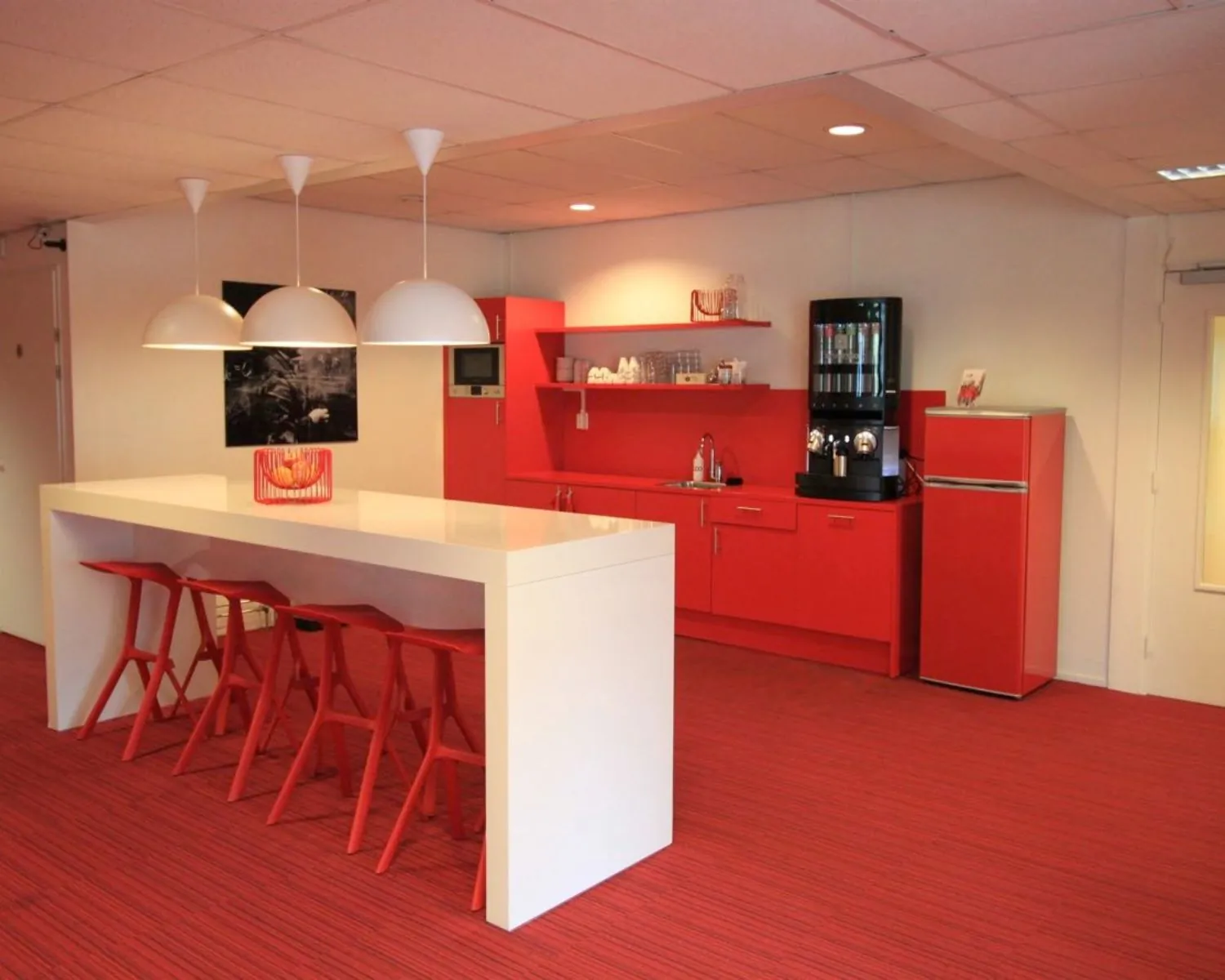 Moderne kantoorkeuken met rood interieur, witte eettafel en barkrukken.