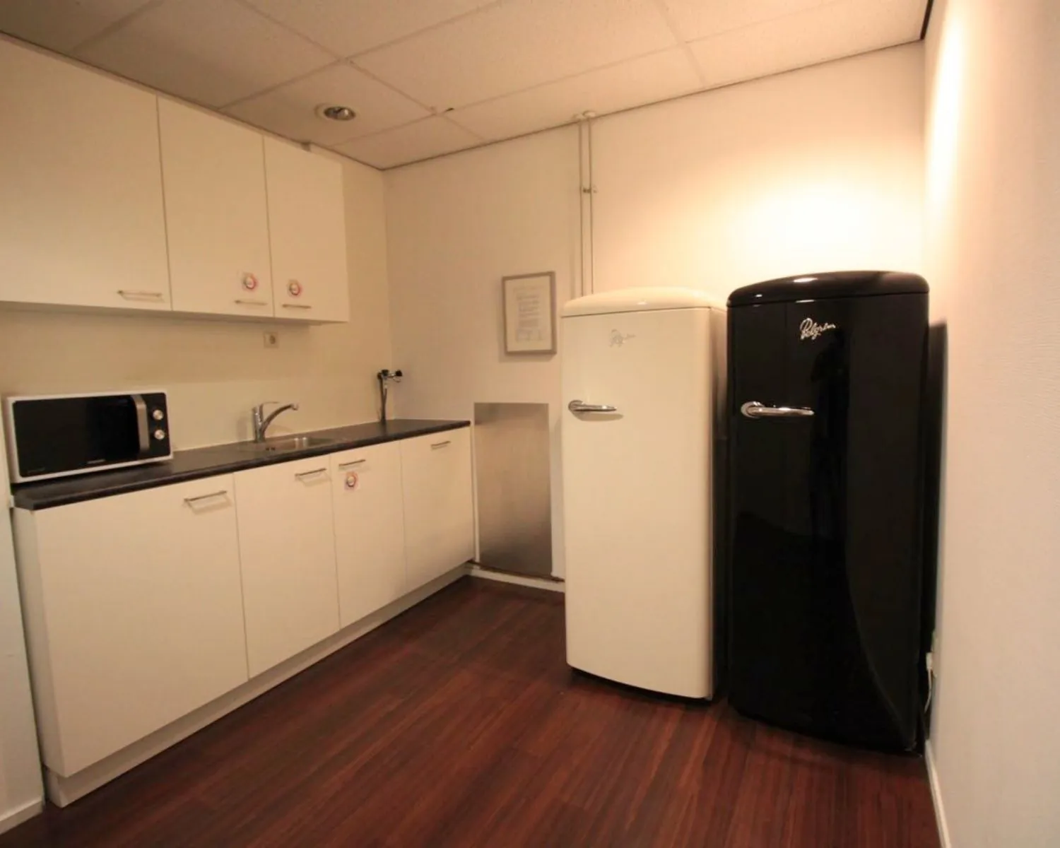 Kleine kitchenette met witte kasten, een magnetron, en twee retro koelkasten in zwart en wit.