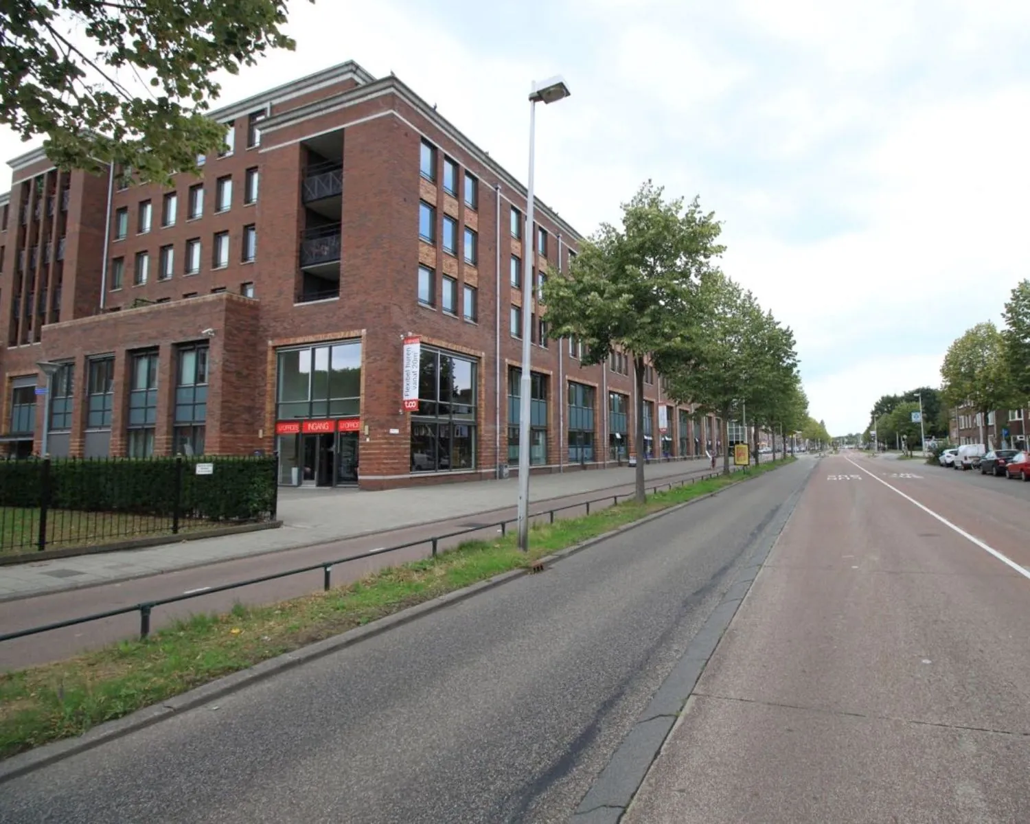 Bakstenen appartementencomplex met winkels op de begane grond langs een brede, boomrijke straat.