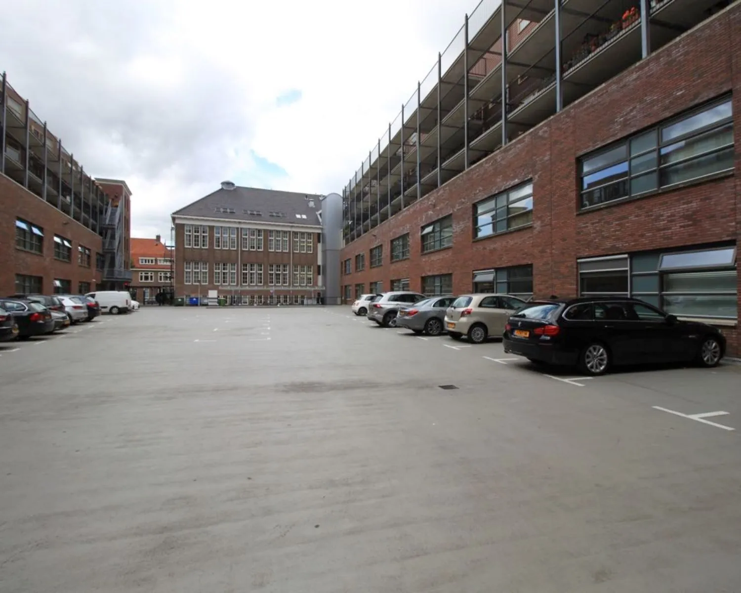 Parkeerplaats omringd door moderne bakstenen appartementsgebouwen en een voormalig schoolgebouw.