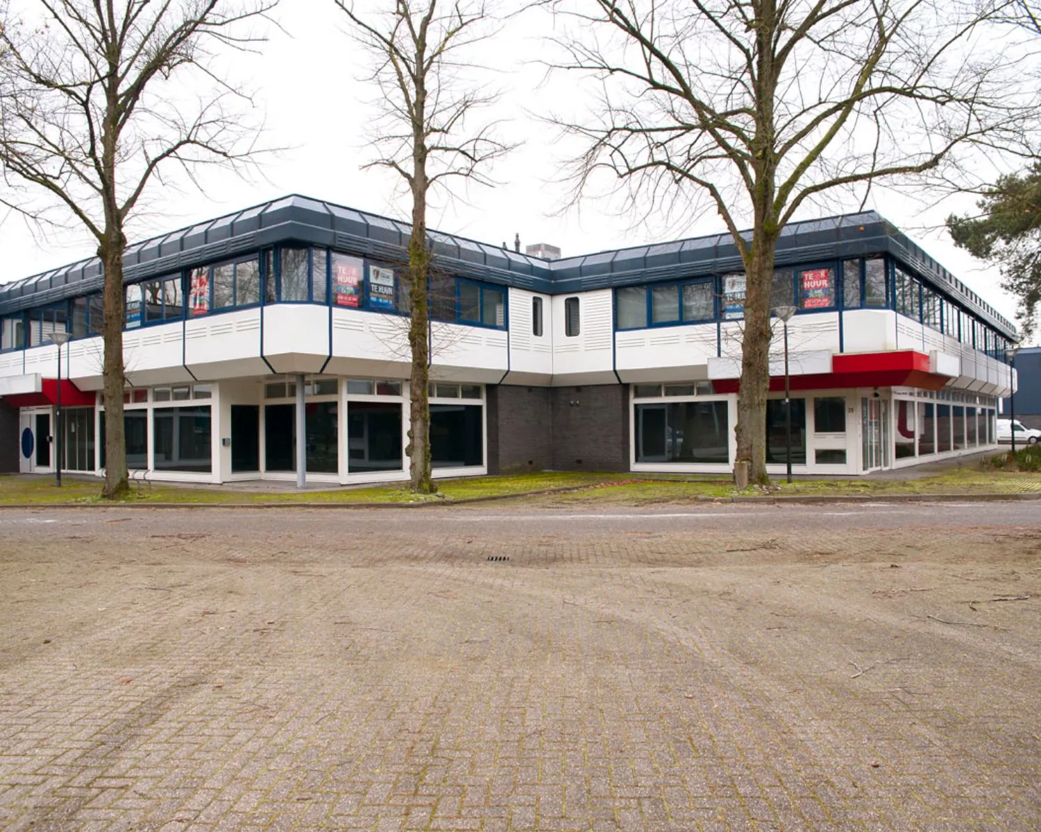 Twee verdiepingen tellend kantoorpand met te huur-borden in de ramen, omringd door bomen en een lege parkeerplaats.