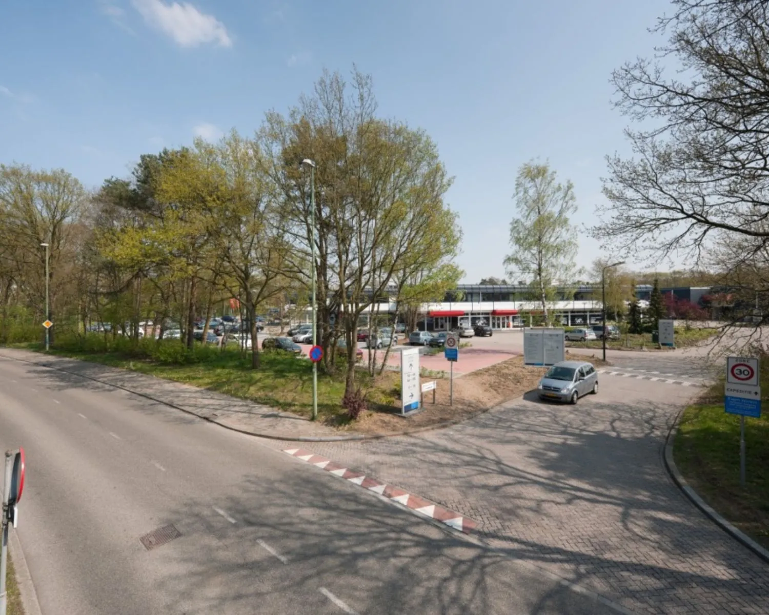 Ingang van een parkeerterrein bij een gebouw, omgeven door bomen en verkeersborden langs een rustige weg.