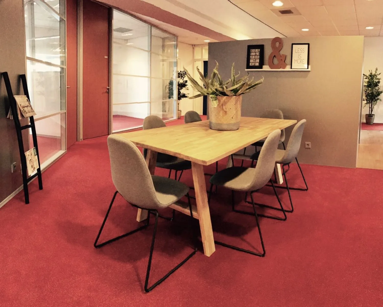 Vergaderruimte met houten tafel, zes grijze stoelen en een grote plant op rood tapijt.
