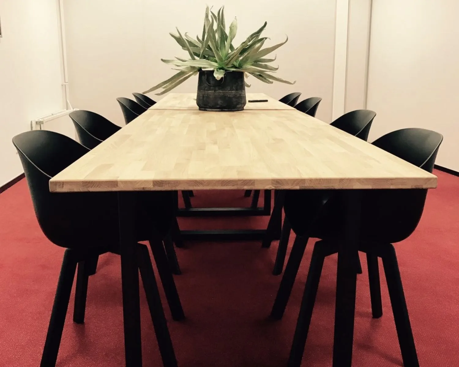 Vergadertafel met houten blad, omringd door zwarte stoelen, op een rode vloer met een groene plant in het midden.