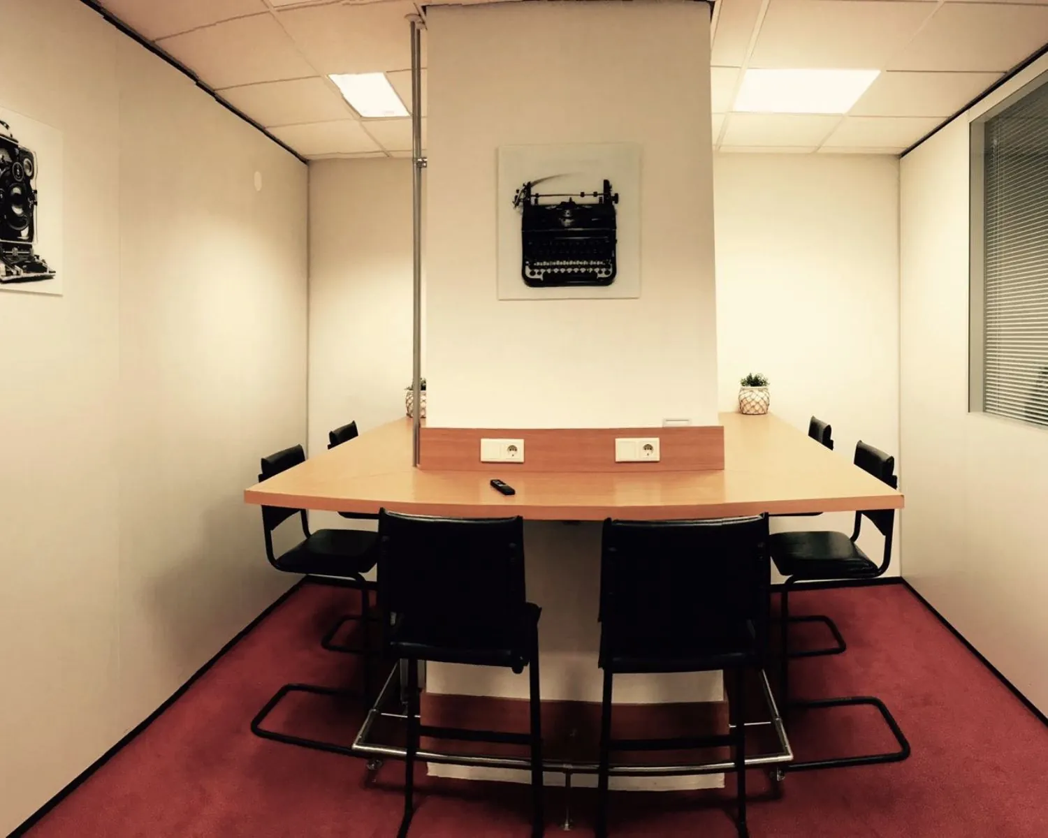 Vergaderruimte met zes zwarte stoelen rond een houten tafel, gescheiden door een muur met stopcontacten en decoraties van typemachinefoto’s aan de wand.