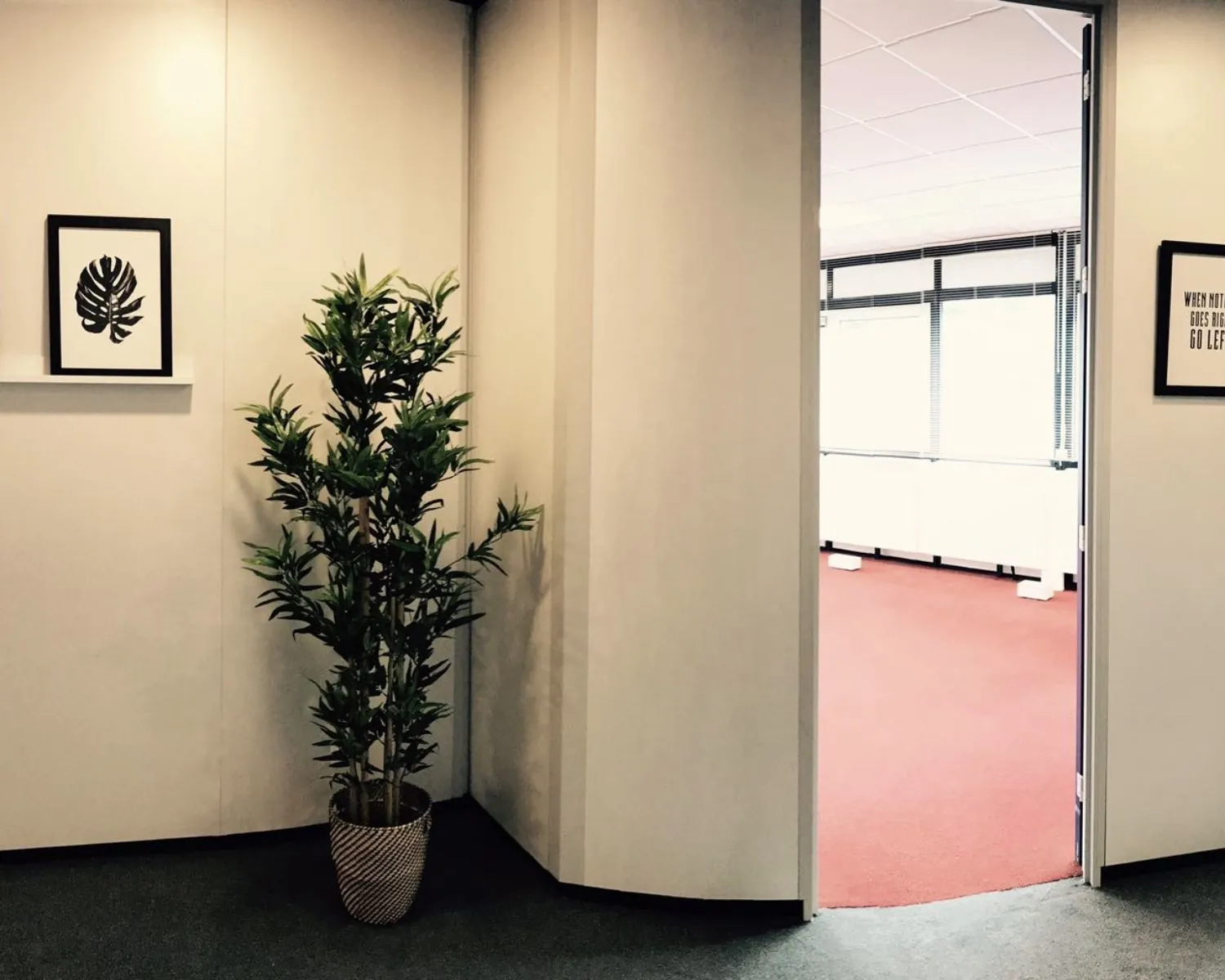 Kantoorhoek met een groene kamerplant, twee ingelijste afbeeldingen aan de muur en een open deur naar een ruimte met rood tapijt.