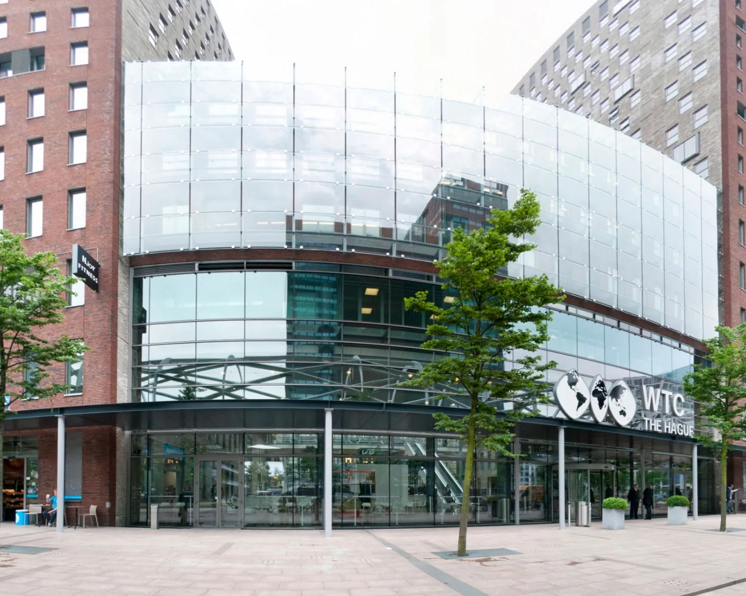 Voorgevel van het WTC The Hague met moderne glazen architectuur en entree aan een plein met jonge bomen.