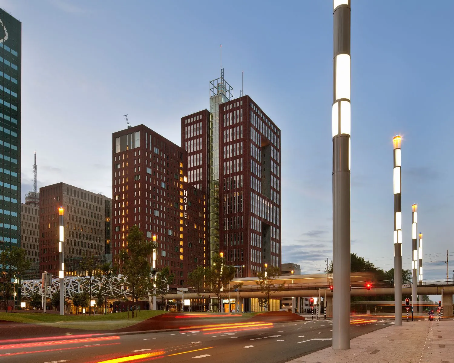 Avondopname van moderne hoogbouw, waaronder het opvallende rode gebouw The Red Apple in Rotterdam, met verlichte lantaarnpalen en lichtstrepen van voorbijrijdend verkeer.